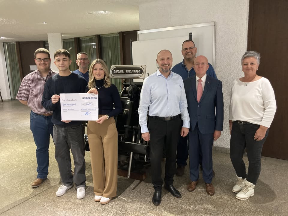 Dieser Alternativtext wurde KI-generiert: Eine Gruppe von Menschen steht in einem Innenraum zusammen und hält einen Scheck. Im Hintergrund befindet sich eine Wand mit einem Schild.