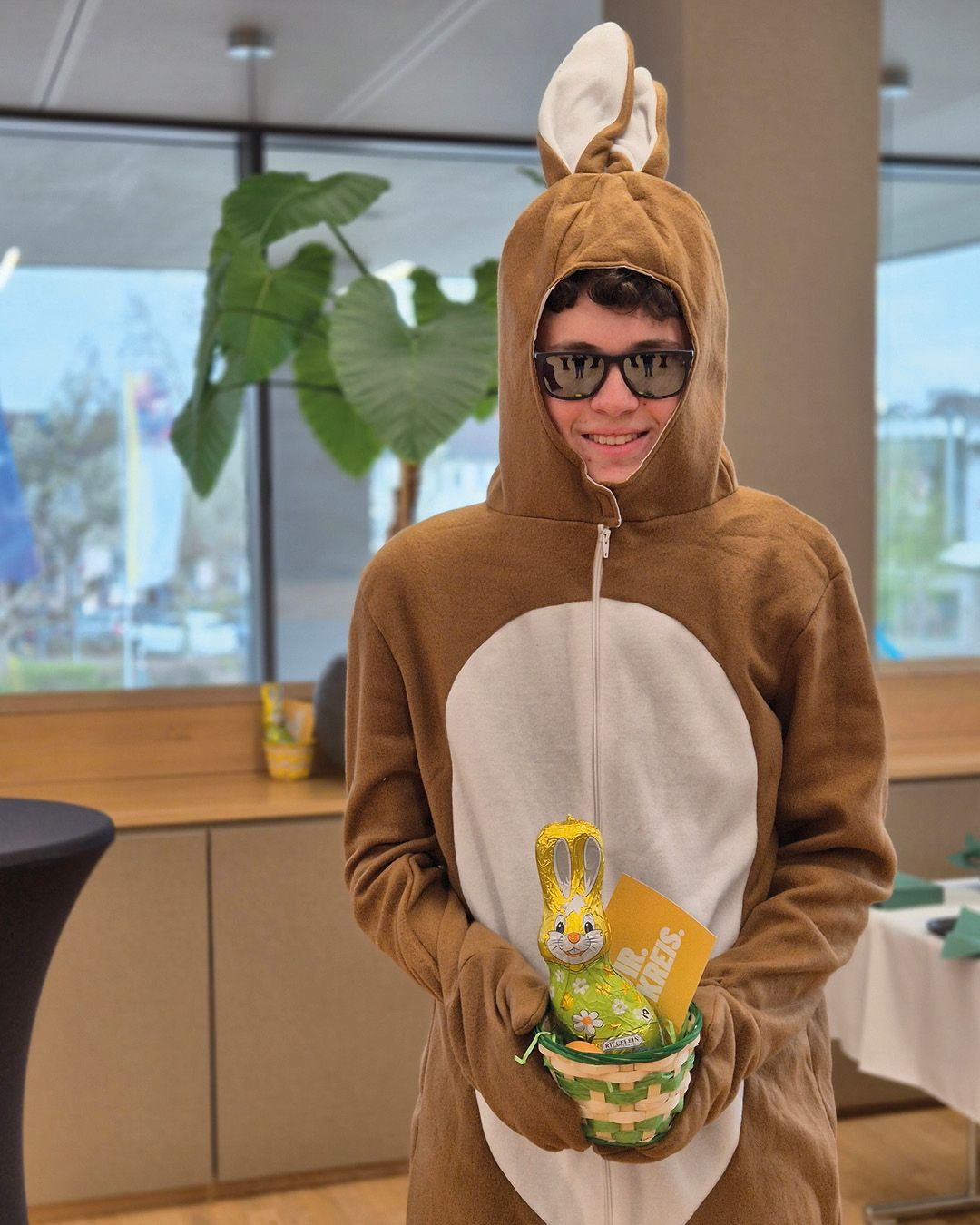 Dieser Alternativtext wurde KI-generiert: Eine Person trägt ein Hasenkostüm und hält einen Osterkorb vor sich. Im Hintergrund sind Pflanzen und ein Fenster zu sehen.
