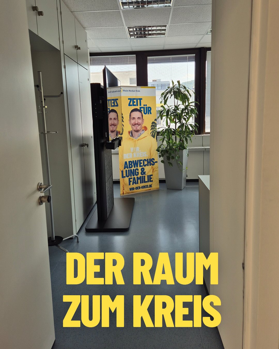 Blick in einen hellen Raum mit einem gelben Standplakat, das eine lächelnde Person in gelbem Hoodie zeigt. Der Text auf dem Plakat lautet „ZEIT FÜR ABWECHSLUNG & FAMILIE“ sowie „WIR. DER KREIS.“. Im Vordergrund steht groß der gelbe Schriftzug „DER RAUM ZUM KREIS“. (KI-Alternativtext)