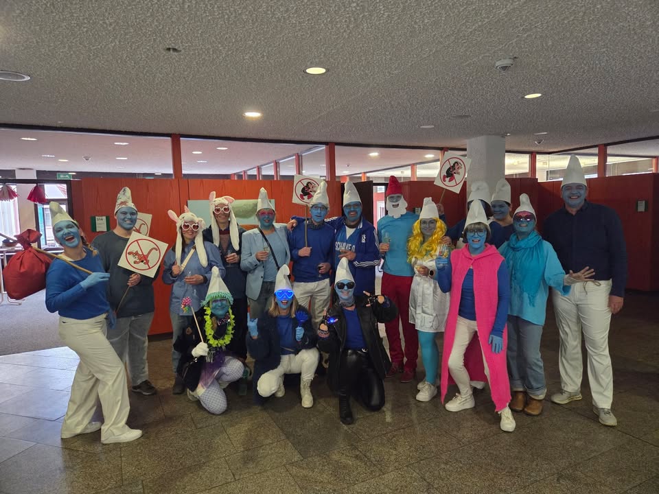 Dieser Alternativtext wurde KI-generiert: Eine Gruppe von Personen in farbenfrohen Kostümen steht in einem Innenraum. Einige der Kostüme ähneln kleinen, blauen Figuren mit weißen Hüten.