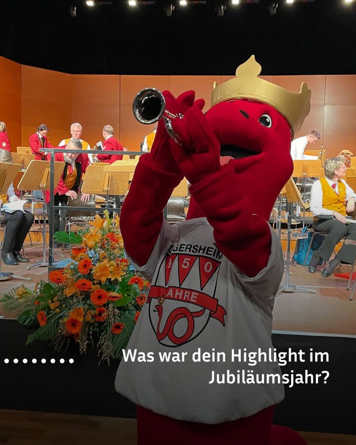 Dieser Alternativtext wurde KI-generiert: Ein Wesen in einem roten Kostüm mit einer Krone spielt in ein Instrument. Im Hintergrund sind Menschen auf einer Bühne und Tische mit Dekorationen zu sehen.