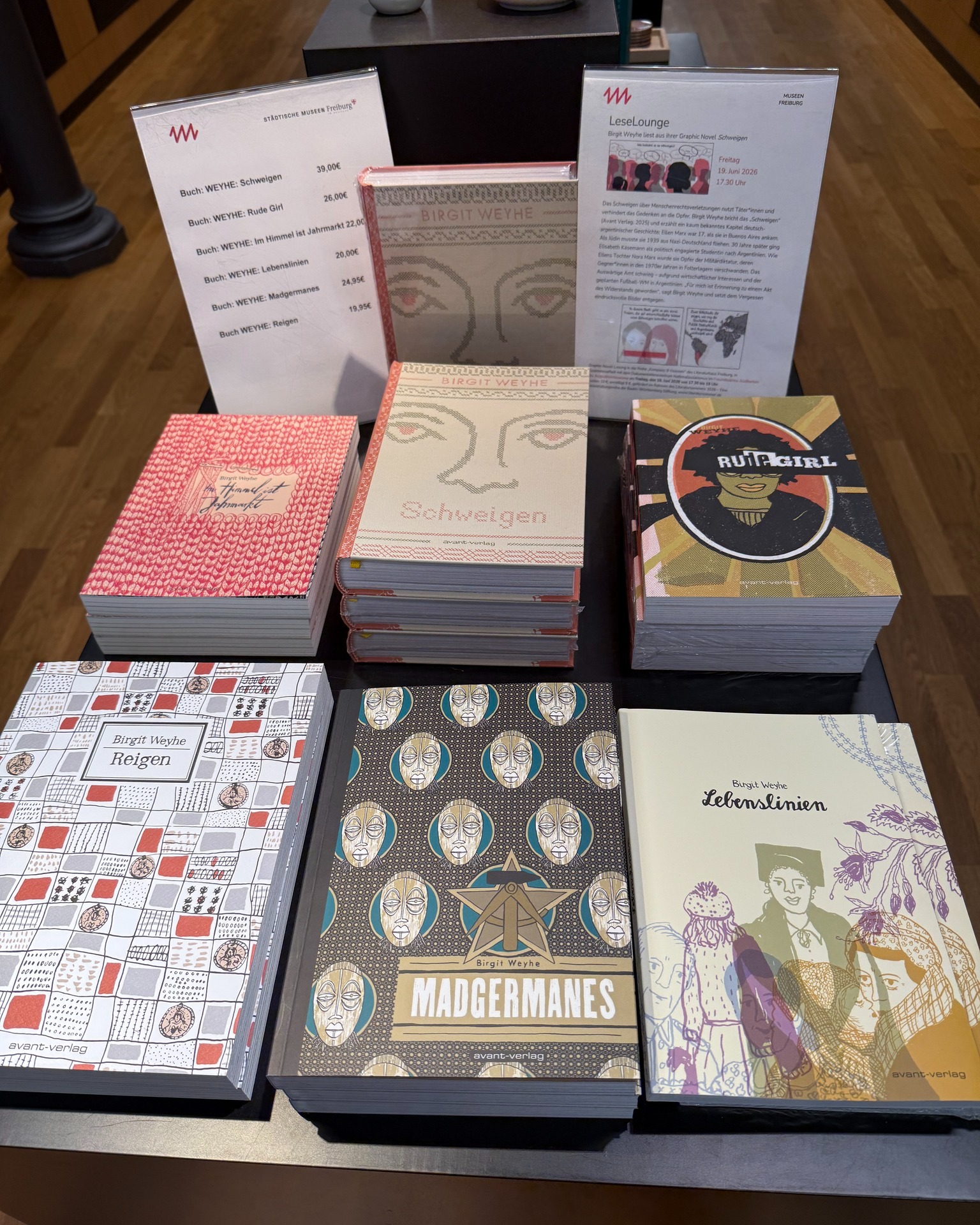 Mehrere Bücher von Birgit Weyhe, darunter „Schweigen“, „Rude Girl“, „Reigen“, „Madgermanes“ und „Lebenslinien“, sind auf einem Tisch gestapelt. Daneben stehen Preisschilder und ein Flyer zur LeseLounge im Städtischen Museum Freiburg. (KI-Alternativtext)