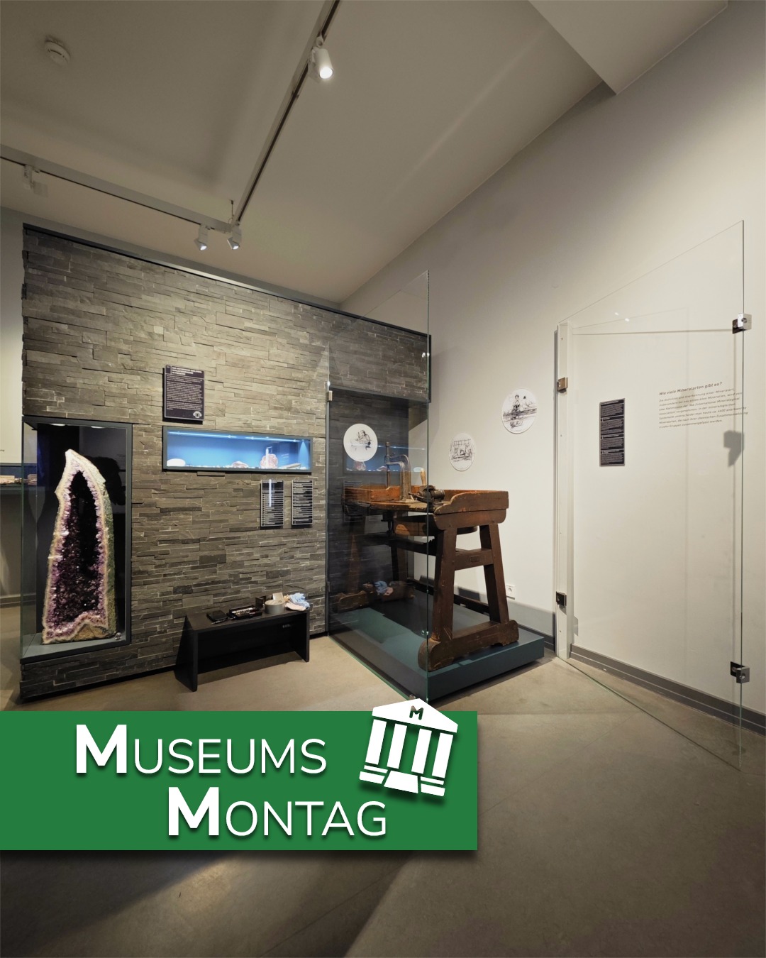 Ein Museumsraum zeigt links eine große Amethyst-Geode in einer Vitrine vor einer Steinwand und in der Mitte einen hölzernen Arbeitstisch in einer Glasschaustelle. Rechts ist eine weiße Wand mit Texttafeln und runden Illustrationen zu sehen. Unten befindet sich ein grüner Banner mit der Aufschrift "MUSEUMS MONTAG" und einem Logo. (KI-Alternativtext)