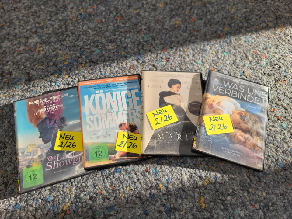 Dieser Alternativtext wurde KI-generiert: Mehrere DVD-Hüllen liegen auf einem türkisfarbenen Teppichboden. Auf den Hüllen sind Filmtitel und Bilder zu sehen, einige haben gelbe Aufkleber mit Zahlen darauf.