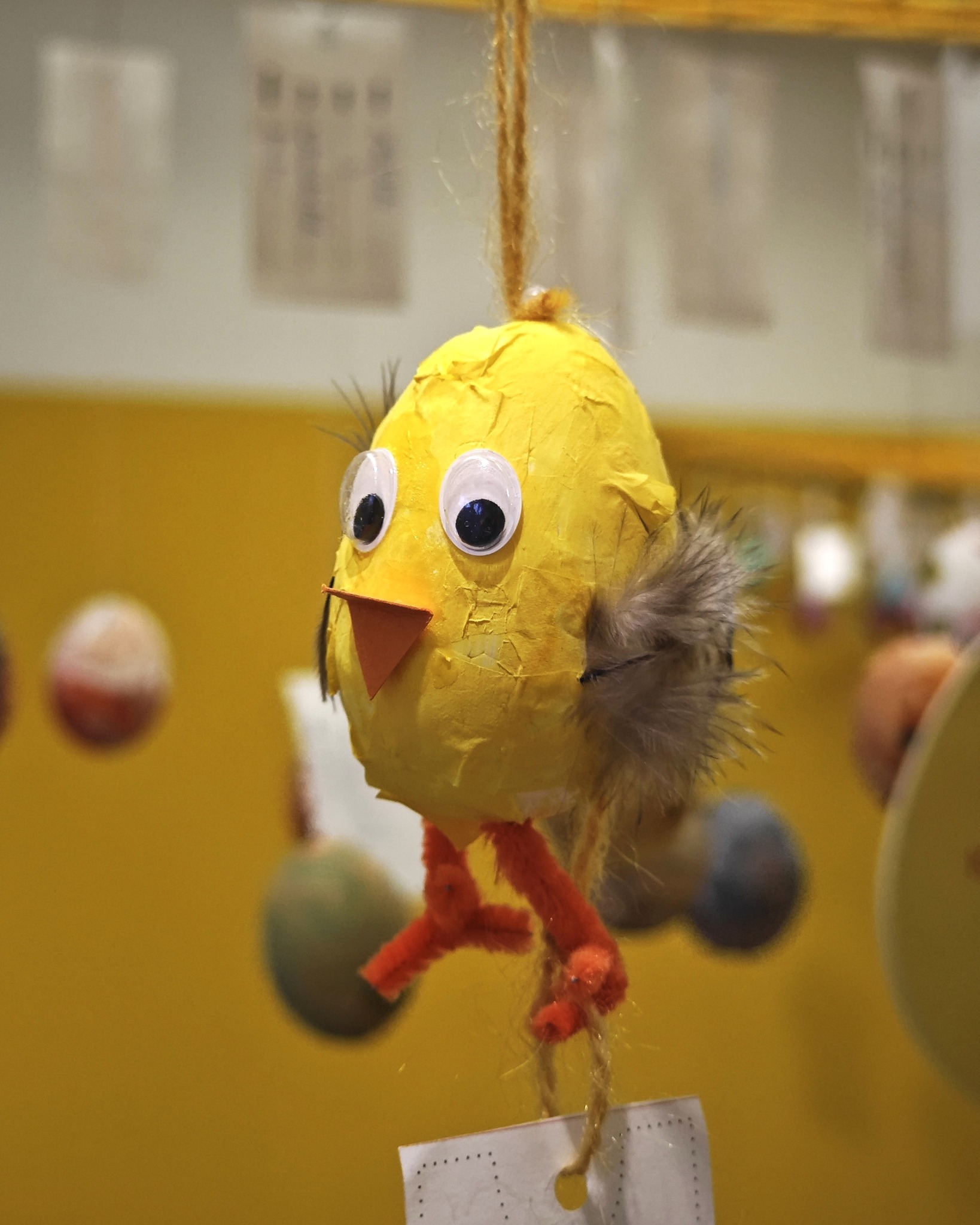 Dieser Alternativtext wurde KI-generiert: Eine gelbe, handgemachte Vogelfigur hängt an einer Schnur. Im Hintergrund sind weitere Dekorationen in verschiedenen Farben zu sehen.