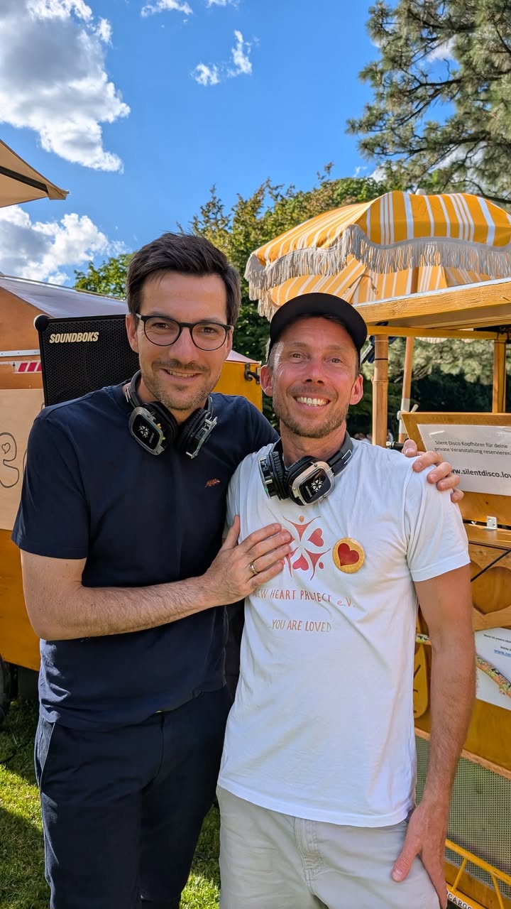 Zwei Personen mit Kopfhörern um den Hals stehen im Freien vor einem gelben Stand mit Sonnenschirm, lächeln in die Kamera. Eine trägt ein weißes T-Shirt mit Herz-Logo und dem Schriftzug „NEW HEART PROJECT e.V. YOU ARE LOVED“. Im Hintergrund sind Bäume und ein blauer Himmel mit Wolken. (KI-Alternativtext)