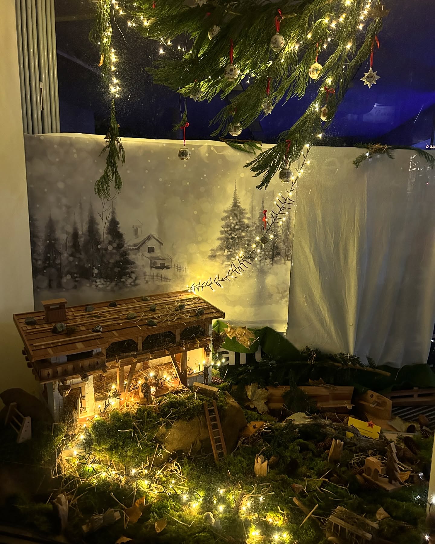 Eine Weihnachtskrippe ist mit Lichtern geschmückt und vor einem Hintergrund mit winterlicher Landschaft platziert. Ein geschmückter Weihnachtsbaum hängt über der Szene.