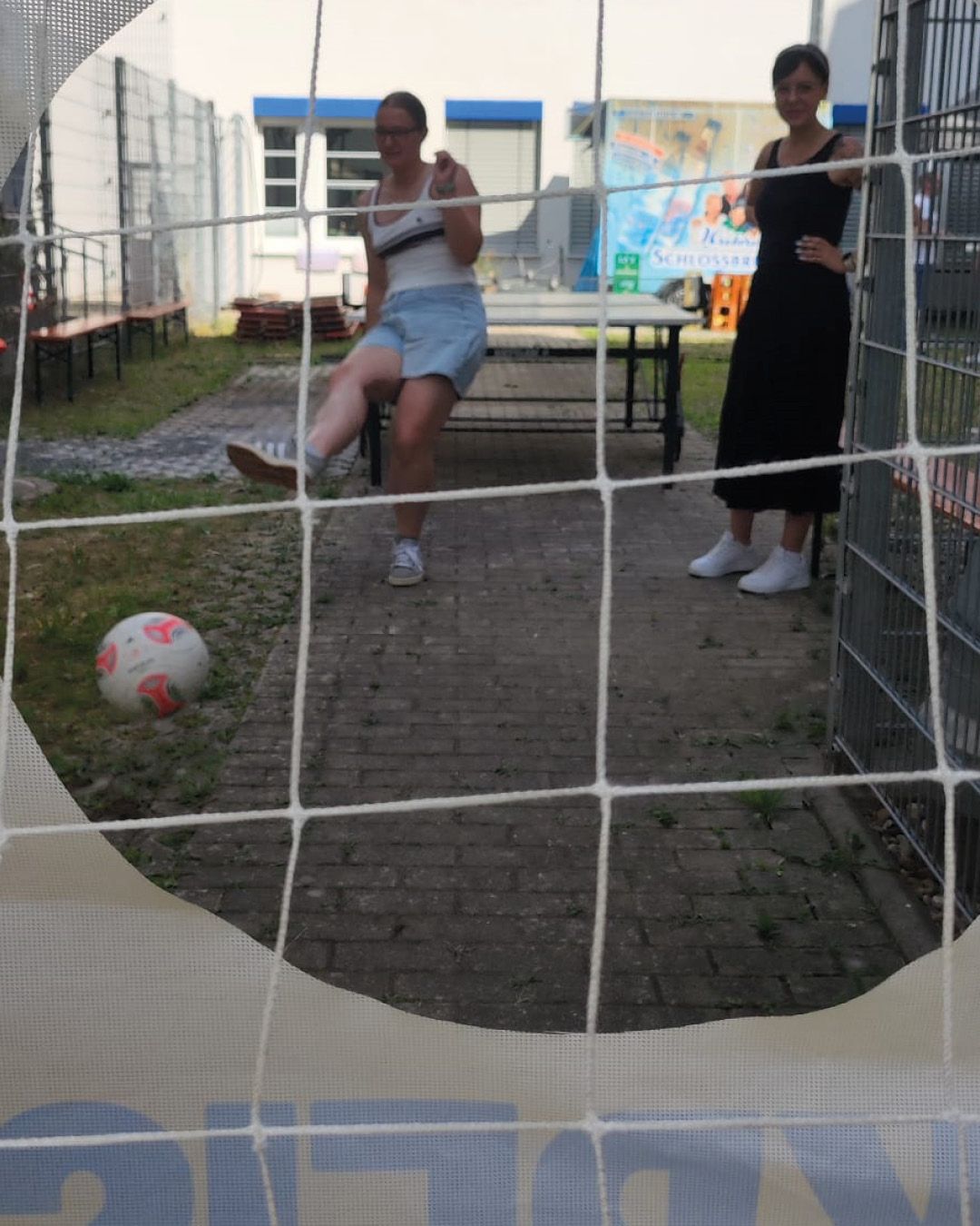 Durch ein weißes Netz im Vordergrund blickt man auf eine Person in Shorts und Tanktop, die einen Fußball auf dem Gras kickt. Rechts daneben steht eine zweite Person in einem langen schwarzen Kleid und beobachtet die Szene. Im Hintergrund stehen eine Tischtennisplatte, Bänke und ein Gebäude mit einem Banner, auf dem teilweise „Schlossbr“ zu lesen ist. (KI-Alternativtext)