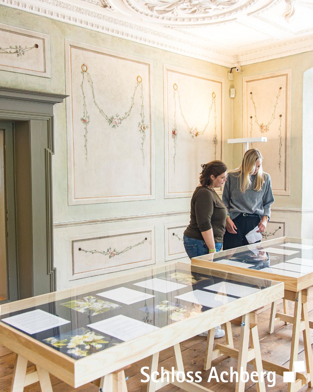 Ein Innenraum mit hellen Wänden und floralen Ornamenten. Zwei Personen betrachten Ausstellungsstücke auf Tischen.