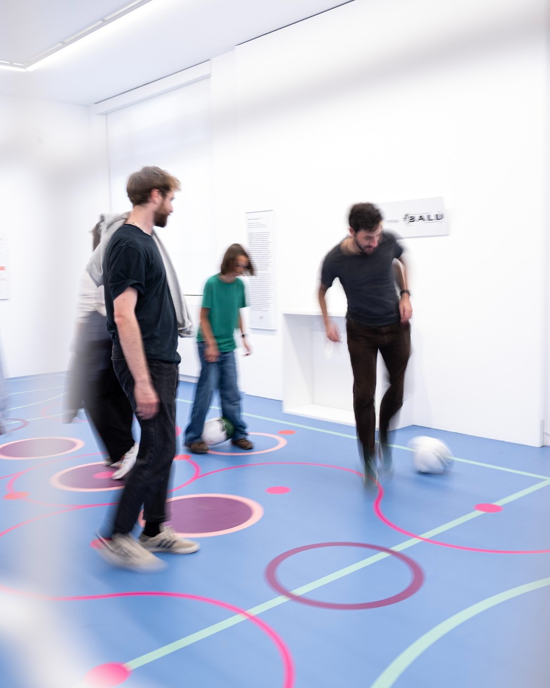Mehrere Personen bewegen sich in einem weißen Raum auf einem blauen Boden, der mit bunten Linien und Kreisen markiert ist. Eine Person auf der rechten Seite kickt einen weißen Ball, während eine weitere Person in der Mitte einen Ball am Fuß hat. An der Wand hängt ein Schild mit der Aufschrift "BALU". (KI-Alternativtext)