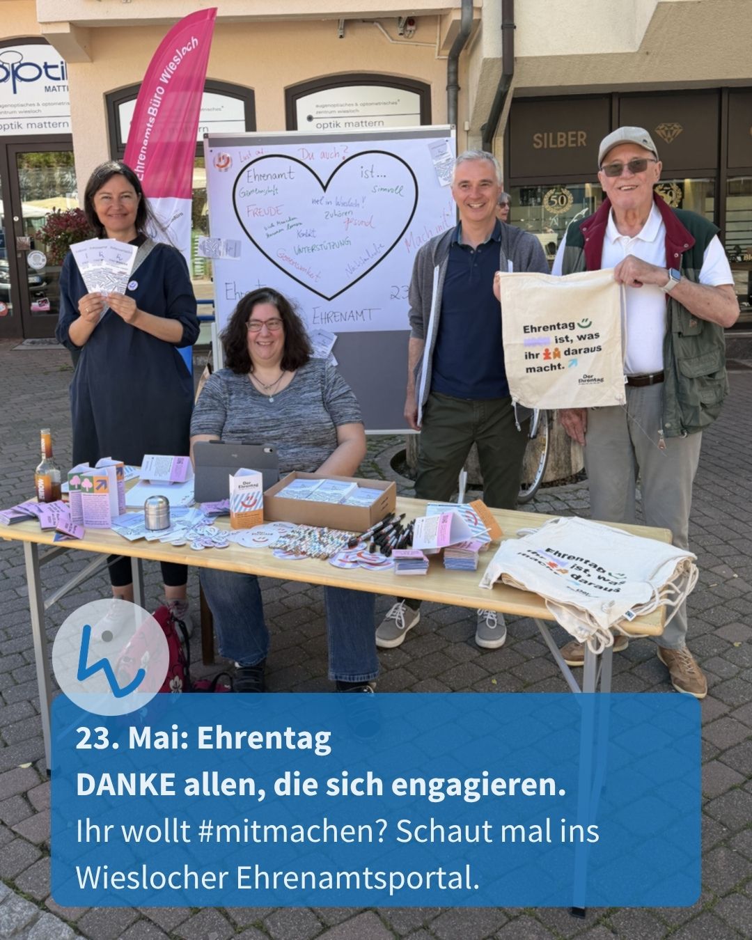 Vier Personen stehen an einem Informationsstand im Freien, einer hält eine Tasche mit der Aufschrift „Ehrentag ist, was ihr daraus macht.“, im Hintergrund ein Whiteboard mit Herz und Texten zum Ehrenamt. Ein Banner nennt „EhrenamtsBüro Wiesloch“. Unten Text: „23. Mai: Ehrentag. DANKE allen, die sich engagieren.“ (KI-Alternativtext)