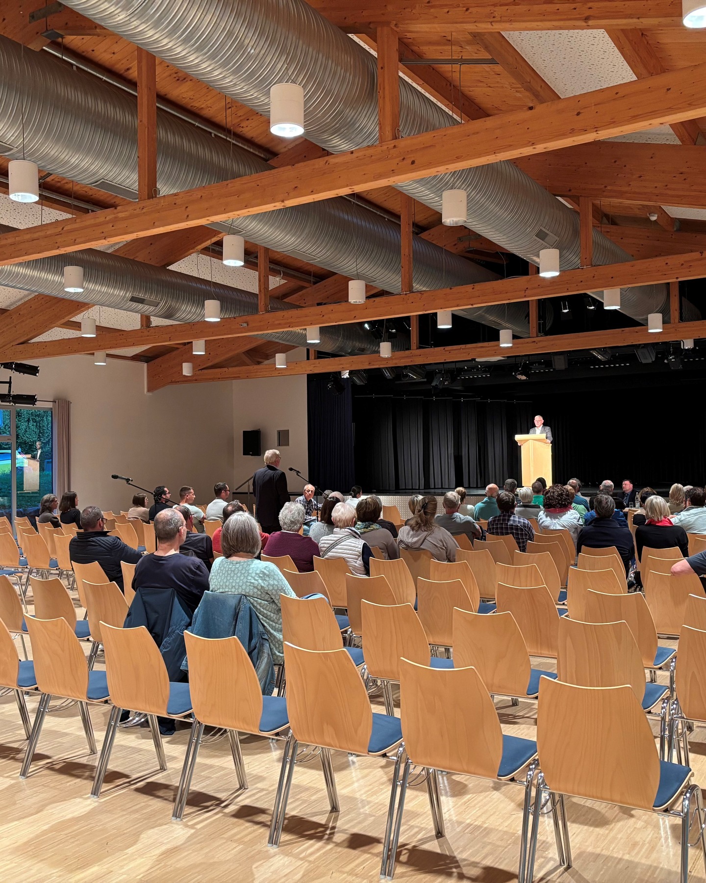 Dieser Alternativtext wurde KI-generiert: Ein Innenraum mit einer Bühne und Reihen von Stühlen, auf denen Menschen sitzen. Im Hintergrund ist ein Fenster zu sehen.