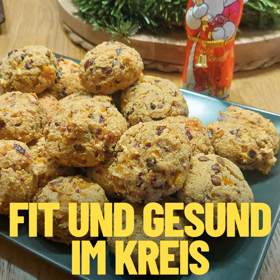 Dieser Alternativtext wurde KI-generiert: Ein Teller mit vielen, runden Keksen, die Früchte enthalten. Im Hintergrund ist ein grüner Zweig und eine rote Figur zu sehen.