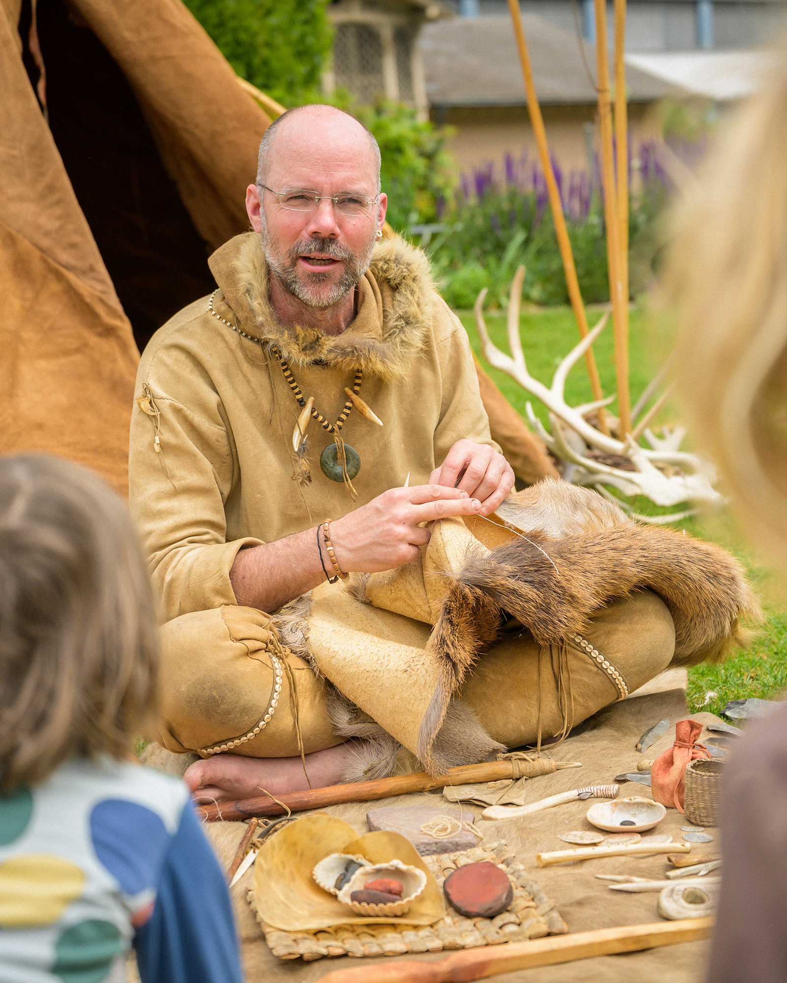 Eine Person in historischer Kleidung sitzt im Freien auf dem Boden, näht mit Nadel und Faden an einem Fell und zeigt dabei auf das Material, während Kinder zuschauen. Um sie herum liegen Werkzeuge, Knochen, Steine und ein Zelt im Hintergrund. (KI-Alternativtext)