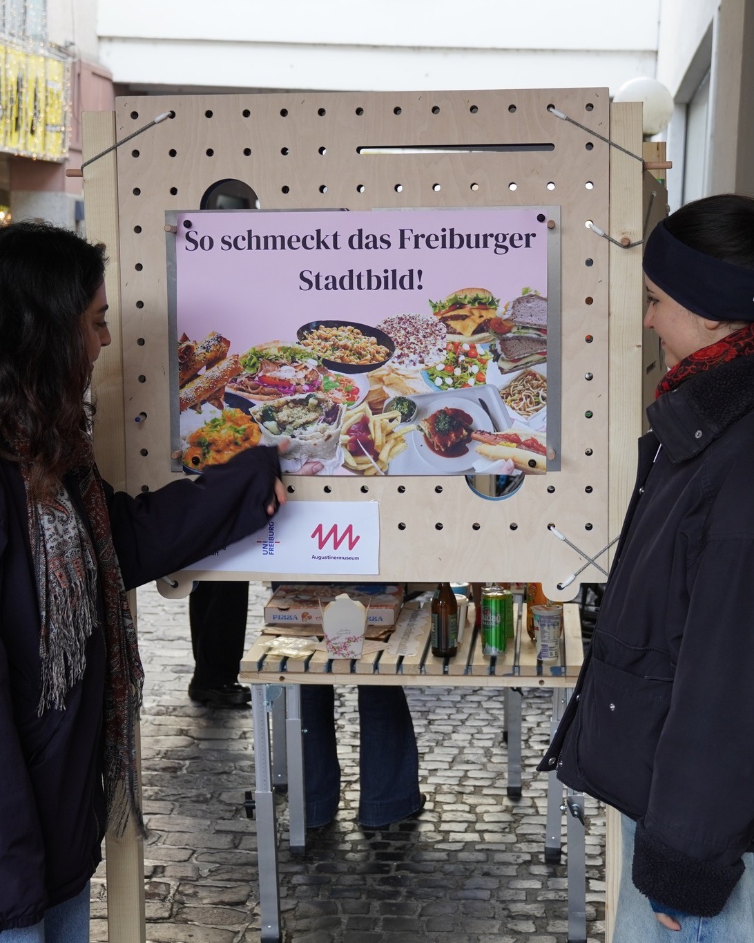 Dieser Alternativtext wurde KI-generiert: Eine Person deutet auf ein Werbeplakat mit verschiedenen Speisen. Darunter steht ein Tisch mit Produkten und Getränken auf einem gepflasterten Platz.