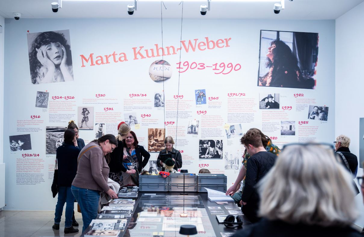 Eine Ausstellung mit vielen Fotos und Texttafeln zeigt das Leben einer Person. Besucher betrachten die Exponate.