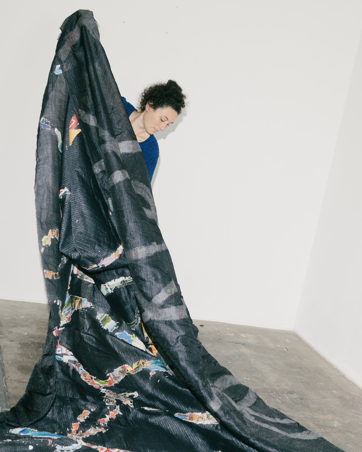 Dieser Alternativtext wurde KI-generiert: Eine Person steht neben einem großen, dunklen Stoffstück, das teilweise auf dem Boden liegt. Das Bild ist farbig und zeigt eine Innenraumansicht.