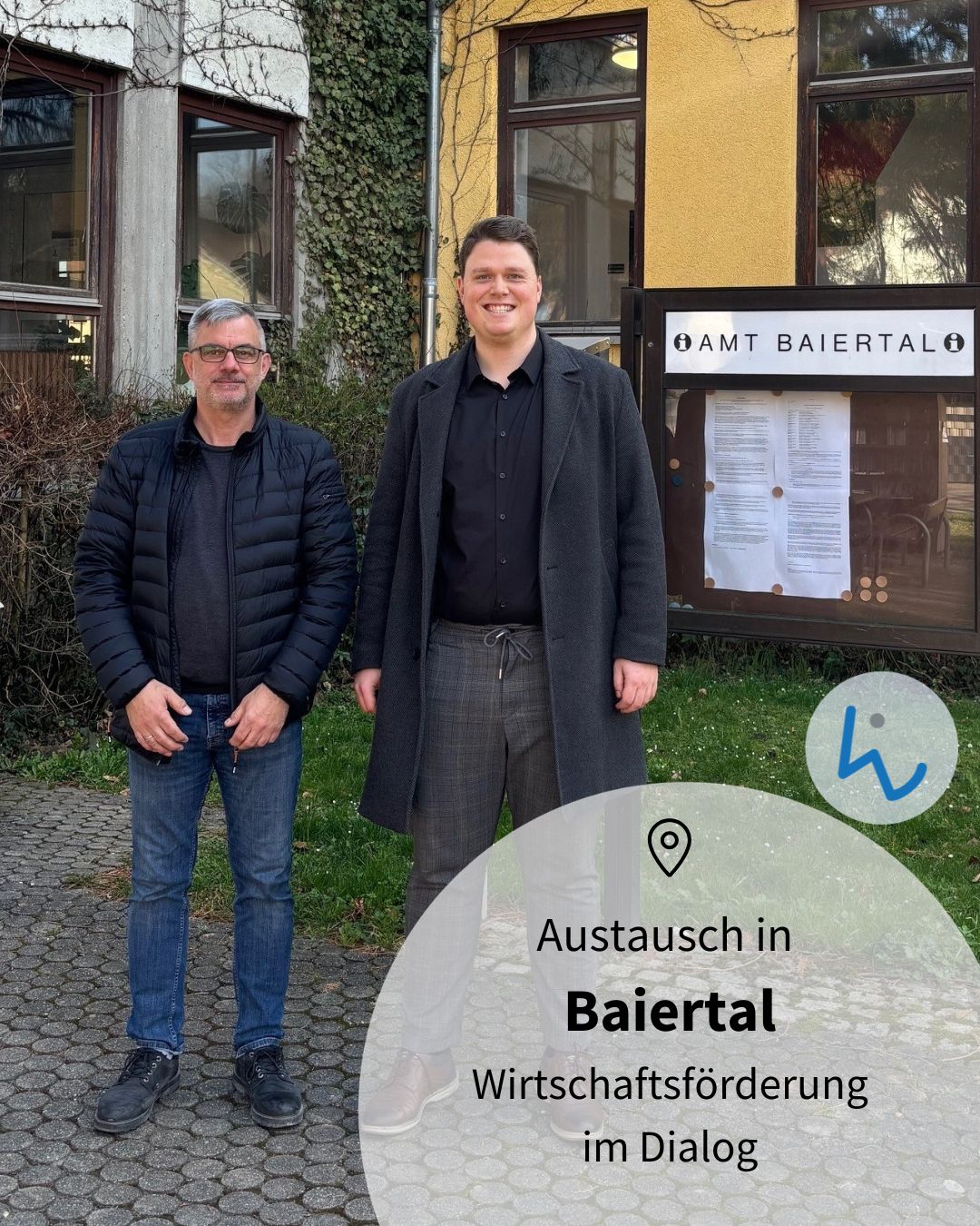 Dieser Alternativtext wurde KI-generiert: Zwei Personen stehen vor einem Gebäude mit einem Informationsbrett. Ein Textfeld mit grafischen Elementen befindet sich im unteren Bildbereich.