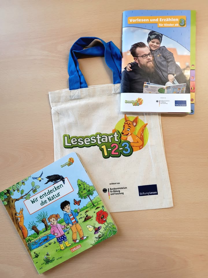 Dieser Alternativtext wurde KI-generiert: Ein Buch, eine Tasche, ein Kleidungsstück und weitere Materialien liegen auf einer hellen Oberfläche. Es handelt sich um Lernmaterialien für Kinder, darunter ein Lesestart-Set.