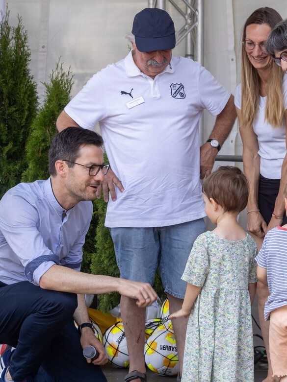 Eine Person kniet links im Bild und hält ein Mikrofon, während sie ein kleines Kind in einem Blumenkleid ansieht. Dahinter steht eine Person in einem weißen Poloshirt und einer Kappe, rechts daneben stehen zwei weitere Personen und ein weiteres Kind. Auf dem Boden liegen zwei gelbe Bälle mit der Aufschrift "DERBYS". (KI-Alternativtext)
