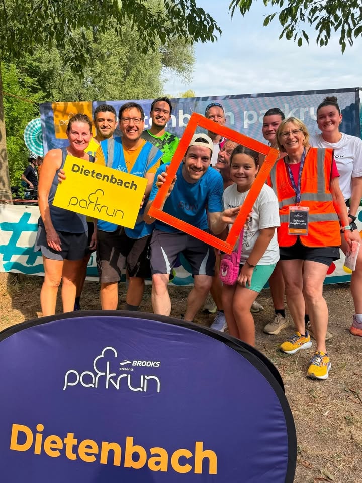 Eine Gruppe von Personen in Sportkleidung posiert im Freien vor einem Banner. Im Vordergrund steht ein blaues Schild mit der Aufschrift "BROOKS presents parkrun Dietenbach". Eine Person hält ein gelbes Schild mit "Dietenbach parkrun", während eine andere einen orangefarbenen Rahmen hält, durch den ein Kind blickt. (KI-Alternativtext)