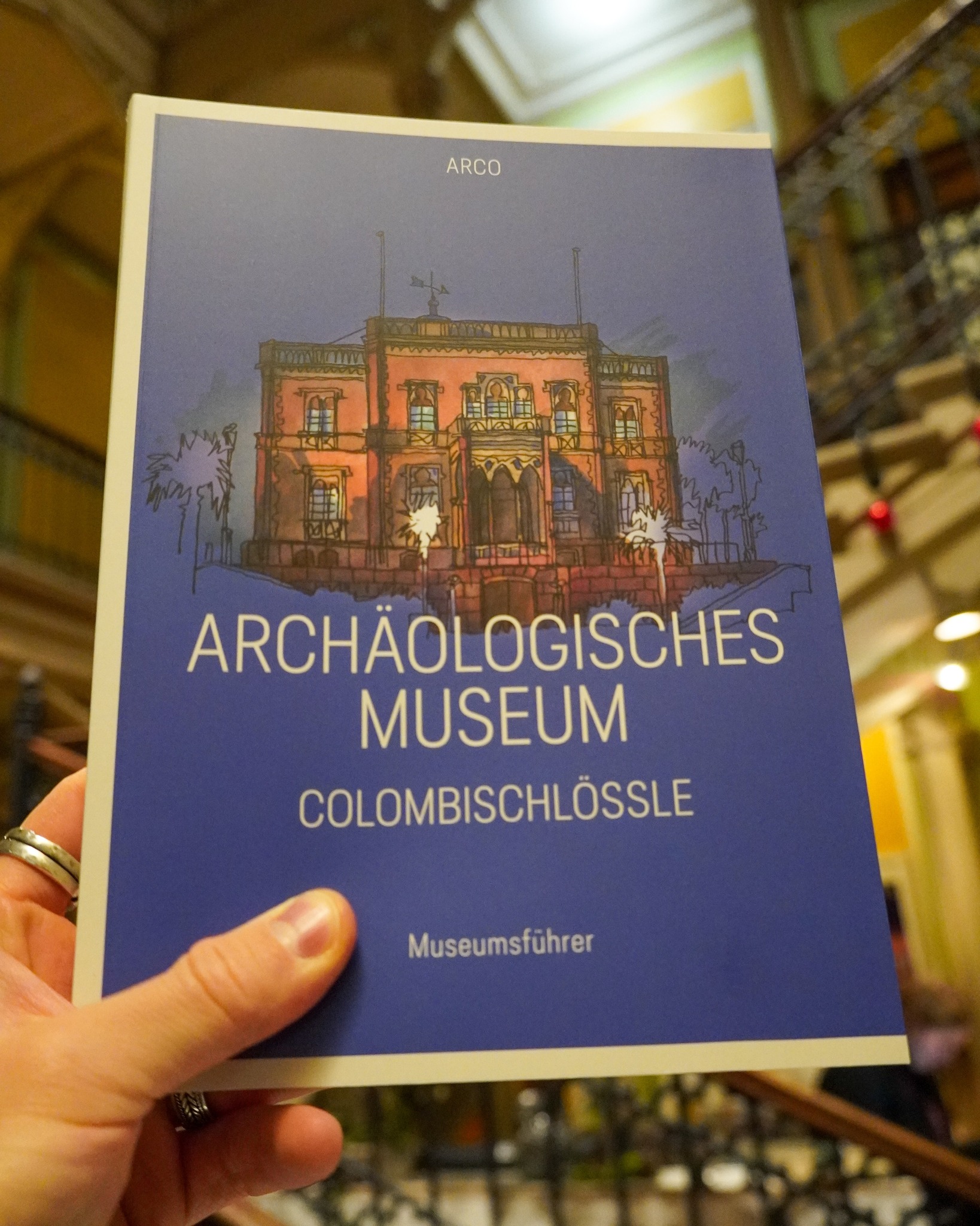 Dieser Alternativtext wurde KI-generiert: Ein Buch liegt auf einer Oberfläche. Das Cover zeigt eine Illustration eines Gebäudes mit dem Titel "Archäologisches Museum Colombischlössle".