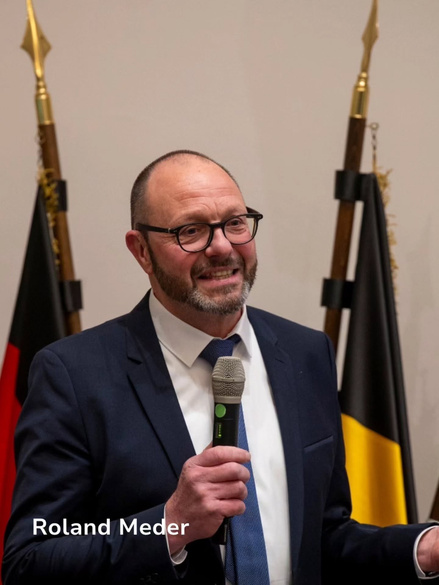 Dieser Alternativtext wurde KI-generiert: Ein Mensch mit Brille spricht in ein Mikrofon, gekleidet in einen Anzug und eine Krawatte. Im Hintergrund sind zwei Flaggen zu sehen.