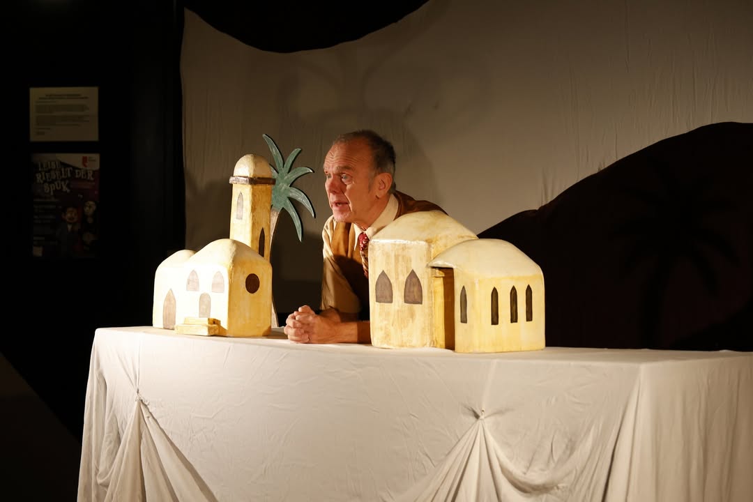 Dieser Alternativtext wurde KI-generiert: Eine Person steht vor einem mit einem Tuch bedeckten Tisch und hält Hände vor Pappmaché-Gebäuden. Im Hintergrund ist ein Plakat zu sehen.