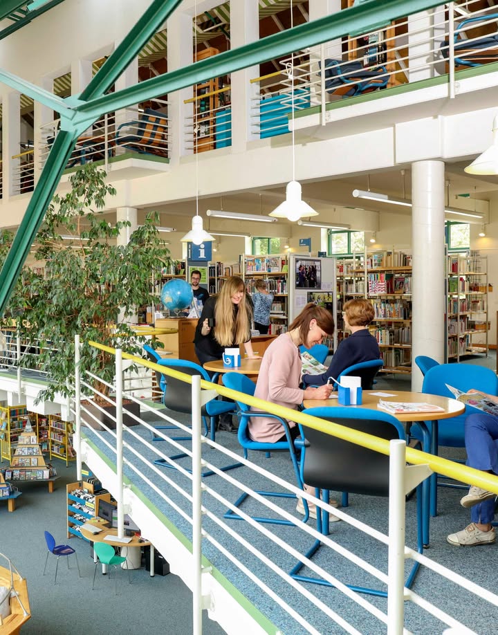 Mehrere Personen sitzen an runden Tischen in einer mehrstöckigen Bibliothek und lesen oder schreiben. Im Hintergrund stehen Bücherregale, ein Globus und eine große Pflanze, während oben ein Balkon mit Geländern verläuft. (KI-Alternativtext)