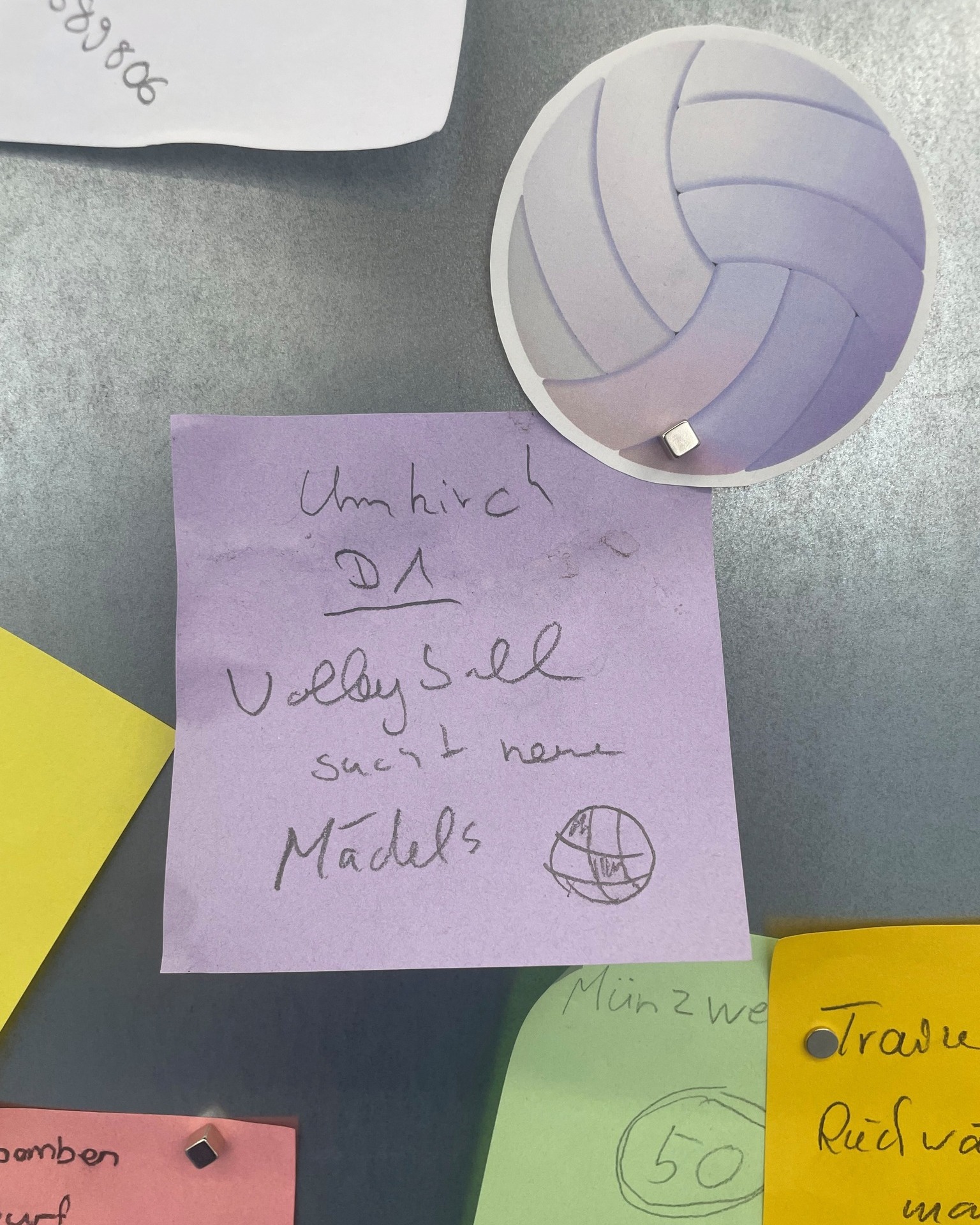 Ein lila Haftnotiz auf einer grauen Pinnwand trägt die handschriftliche Aufschrift „Umhirsch DA Volleyball sucht neue Mädels“ sowie eine kleine Skizze eines Balls. Direkt darüber ist ein rundes Bild eines Volleyballs befestigt. Im Hintergrund sind Teile weiterer bunter Notizzettel mit handschriftlichen Notizen zu erkennen. (KI-Alternativtext)