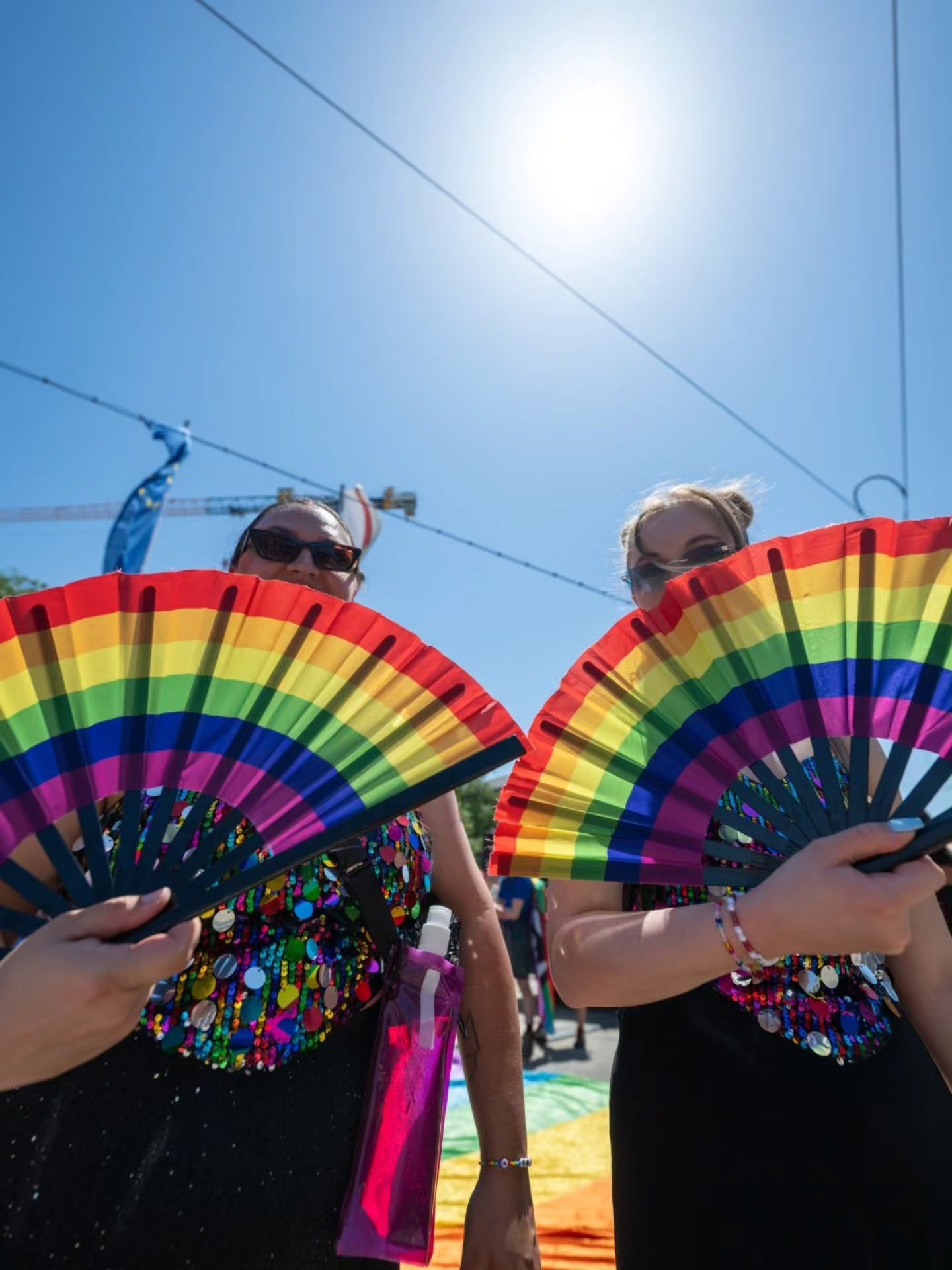 Zwei Personen halten bunte Regenbogenfächer vor sich, tragen glitzernde Oberteile und Sonnenbrillen, im Hintergrund strahlt die Sonne an einem klaren blauen Himmel. (KI-Alternativtext)