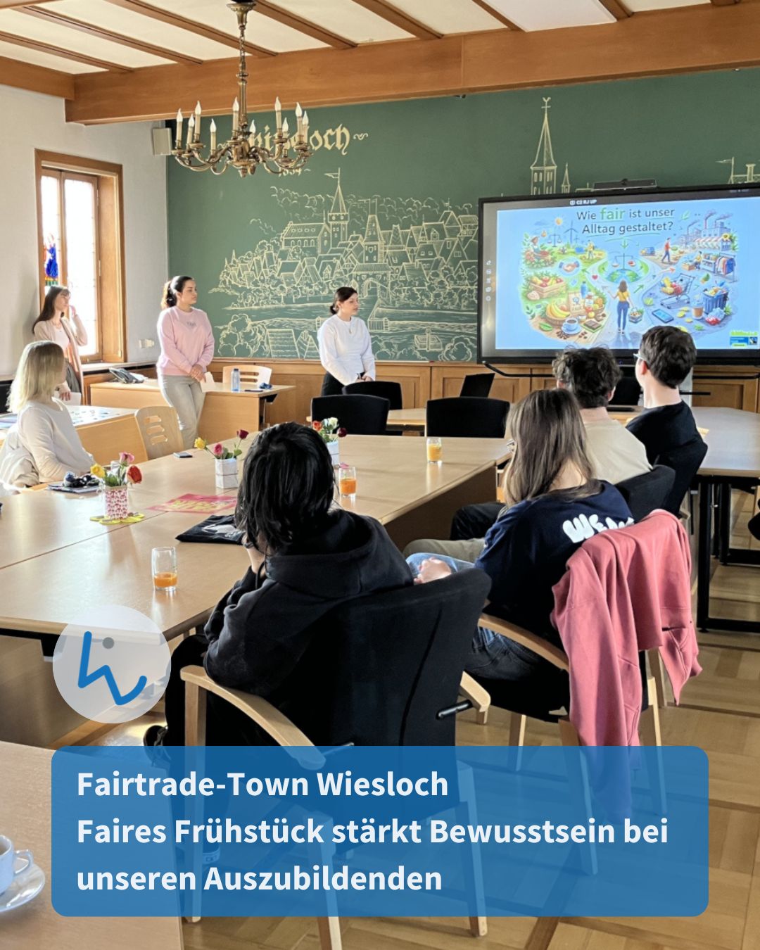 Dieser Alternativtext wurde KI-generiert: Eine Gruppe von Menschen sitzt um einen großen Tisch in einem hellen Raum. Eine Präsentation mit einer farbenfrohen Grafik ist im Hintergrund zu sehen.