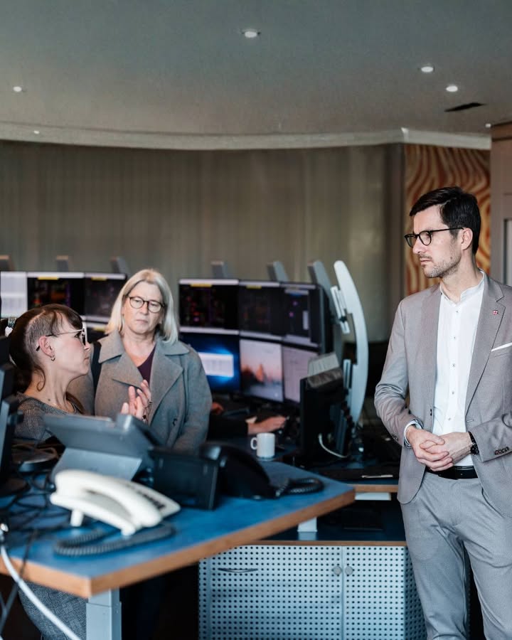 Dieser Alternativtext wurde KI-generiert: Mehrere Personen arbeiten an Computern in einem modernen Büro. Eine Person in einem Anzug steht abseits und blickt in die Runde.