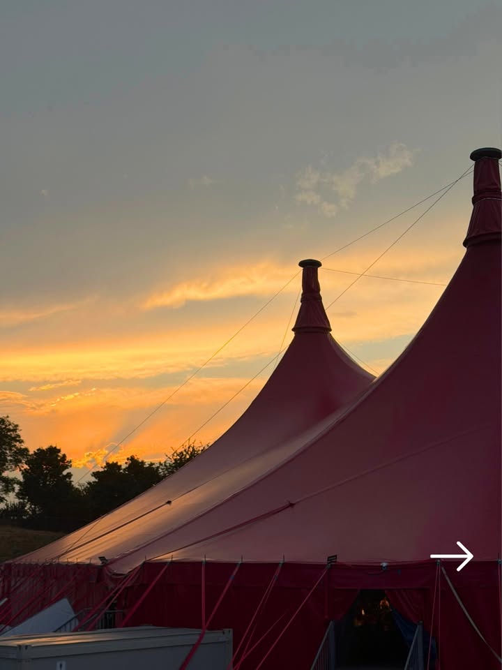 Zwei rote Zelte mit spitzen Dächern stehen vor einem orange-gelben Abendhimmel. Ein weißer Pfeil im unteren rechten Bildrand zeigt nach rechts. (KI-Alternativtext)