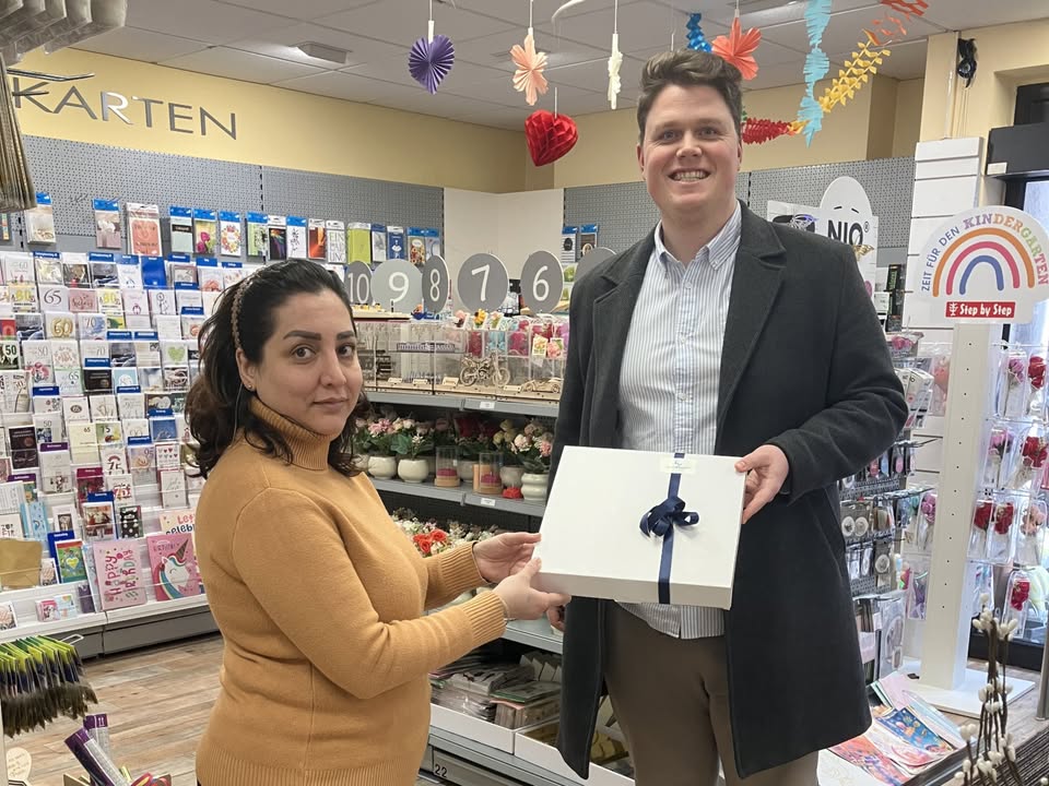 Dieser Alternativtext wurde KI-generiert: Eine Person überreicht einer anderen eine Geschenkbox mit Schleife in einem Geschäft. Im Hintergrund sind Karten, Blumen und Dekorationen zu sehen.