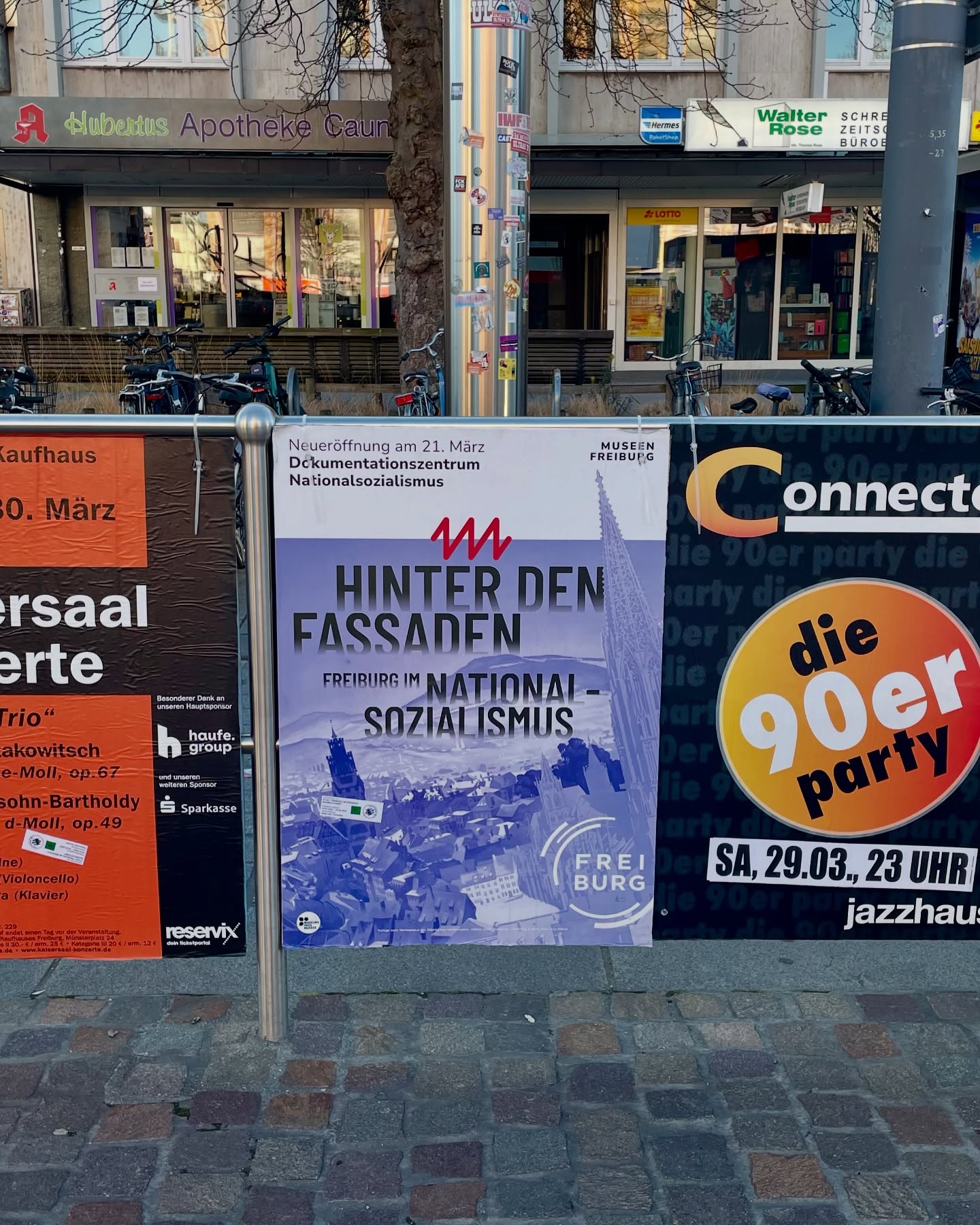 Drei Plakate vor einem Gebäude zeigen ein Konzert, eine Ausstellungseröffnung und den Hinweis auf ein Dokumentationszentrum.  Die Plakate stehen an einem Zaun vor einem Gehweg.