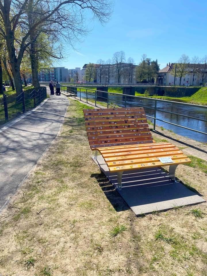 Dieser Alternativtext wurde KI-generiert: Ein Parkweg entlang eines Flusses mit Bäumen und einer Bank im Vordergrund. Die Aufnahme ist farbig und zeigt eine sonnige Szene.