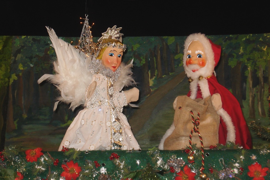 Zwei Puppen, eine im Engelskostüm, die andere im Weihnachtsmannkostüm, stehen auf einer Bühne vor einem Waldhintergrund.