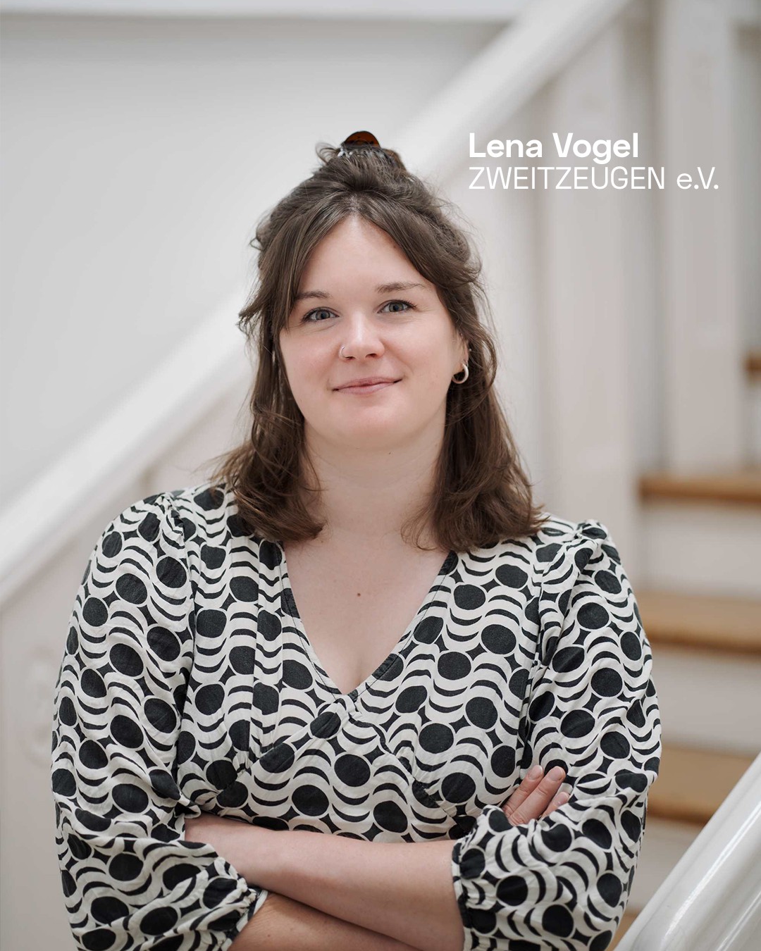 Eine Person mit schulterlangen braunen Haaren und einem weiß-schwarz gemusterten Oberteil steht mit verschränkten Armen vor einer Treppe. Rechts oben steht der Text „Lena Vogel ZWEITZEUGEN e.V.“. (KI-Alternativtext)
