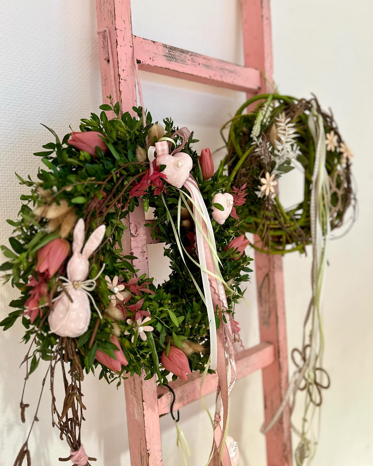 Zwei Kränze hängen an einer rosafarbenen Leiter. Der eine Kranz ist mit Blüten, Schleifen und einem Hasen verziert.
