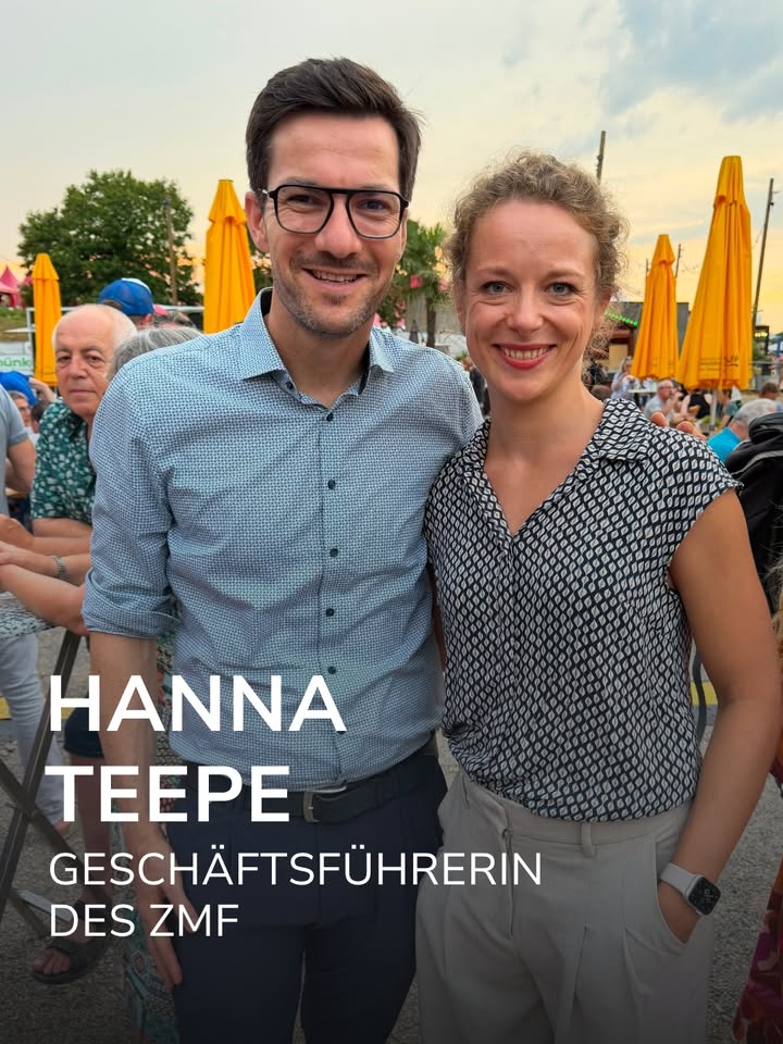 Zwei Personen stehen nebeneinander im Freien und lächeln in die Kamera. Die Person links trägt eine Brille und ein hellblaues Hemd, die Person rechts ein schwarz-weiß gemustertes ärmelloses Oberteil. Im Hintergrund sind gelbe Sonnenschirme und weitere Personen zu sehen. Ein Texteinblendung lautet: „HANNA TEEPE GESCHÄFTSFÜHRERIN DES ZMF“. (KI-Alternativtext)
