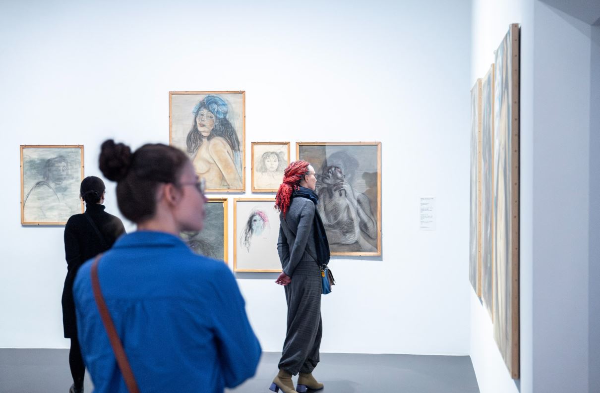 Menschen betrachten in einer Galerie Kunstwerke an den Wänden. Die Bilder zeigen Porträts und Zeichnungen.