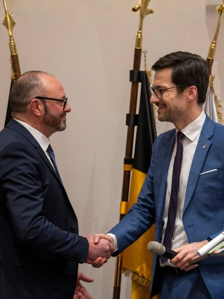 Dieser Alternativtext wurde KI-generiert: Zwei Personen in Anzügen geben sich die Hand vor einer Fahne und goldenen Standarten. Das Bild ist in Farbe gehalten.