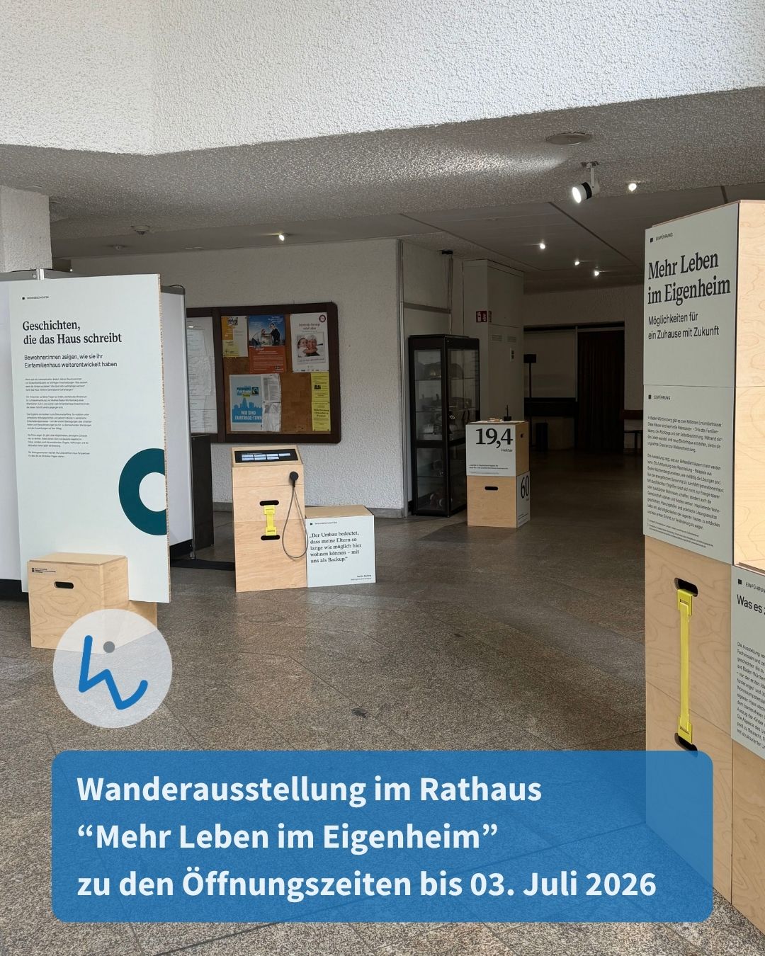 Wanderausstellung im Rathaus mit Informationsständen und Holzkisten, die Zahlen wie „19,4“ und „60“ zeigen. Ein Schild trägt den Titel „Mehr Leben im Eigenheim“. Die Ausstellung läuft bis 03. Juli 2026. (KI-Alternativtext)