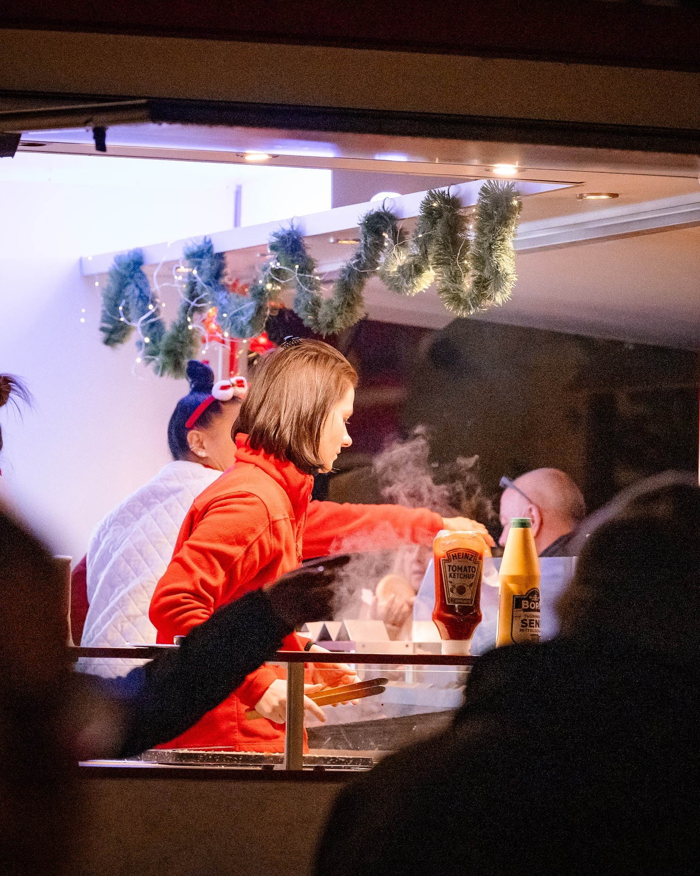 Eine Person in roter Jacke bereitet Essen an einem Marktstand zu. Es gibt Dampf und Flaschen mit Ketchup und Senf.