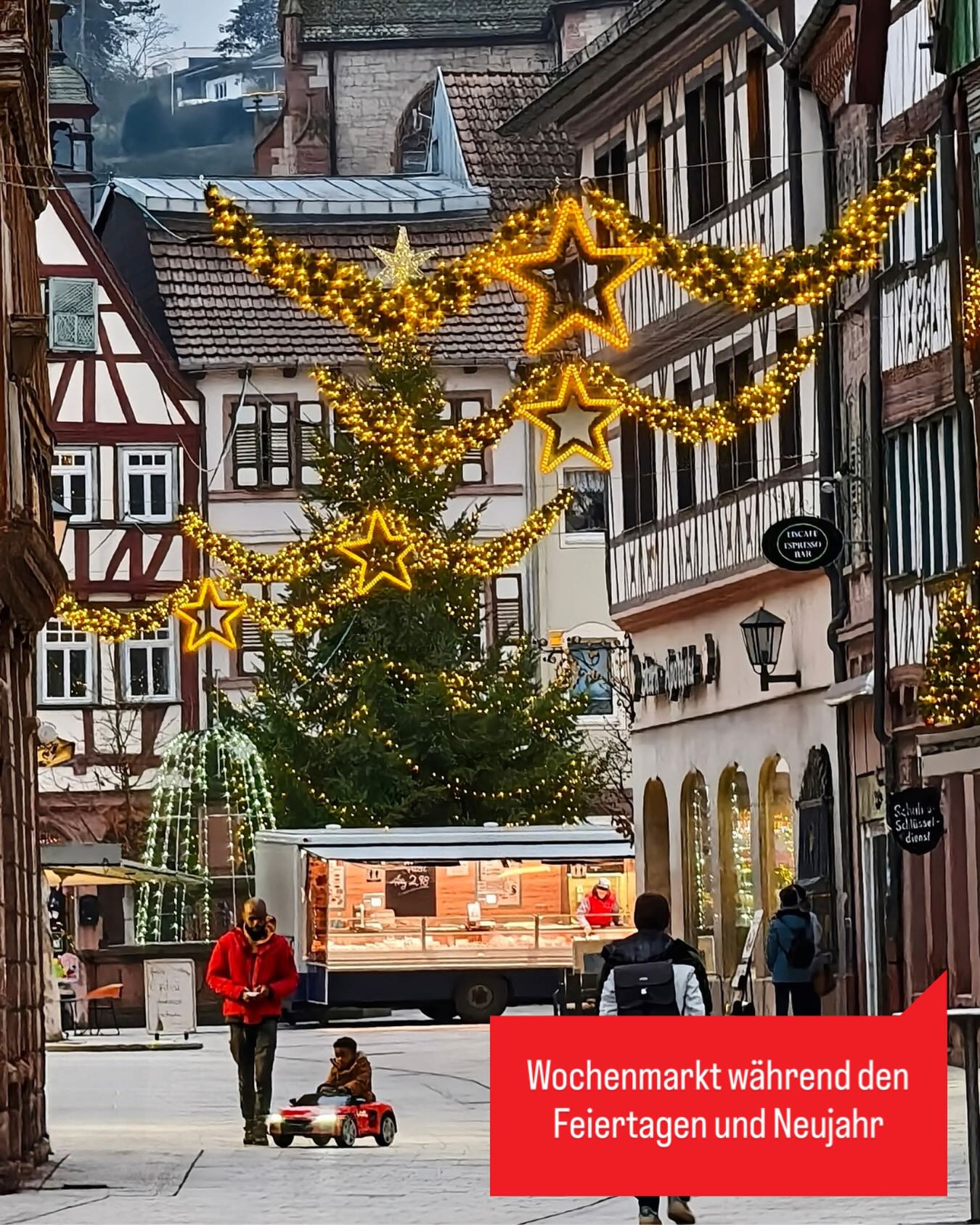 Dieser Alternativtext wurde KI-generiert: Ein Blick auf eine stimmungsvolle Gasse mit Fachwerkhäusern, geschmückt mit Weihnachtsbeleuchtung. Im Vordergrund stehen Menschen und ein kleiner Wagen.