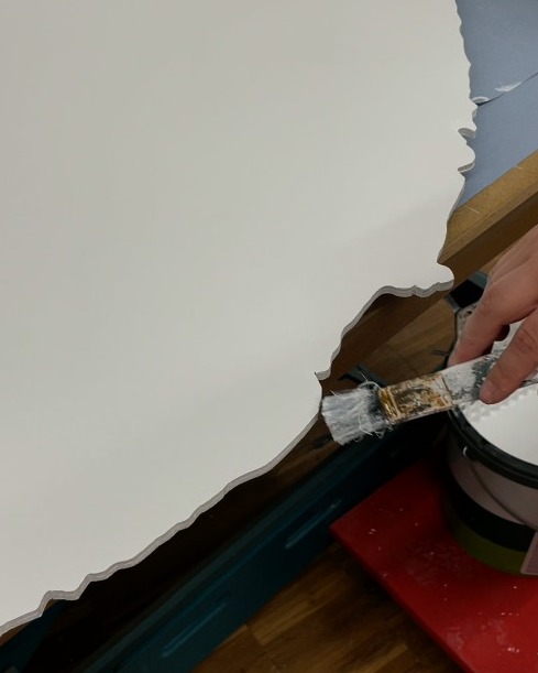 Dieser Alternativtext wurde KI-generiert: Ein Pinsel wird benutzt, um eine Wand weiß zu streichen. Im Hintergrund ist ein Teil eines Holzrahmens und ein Farbeimer zu sehen.