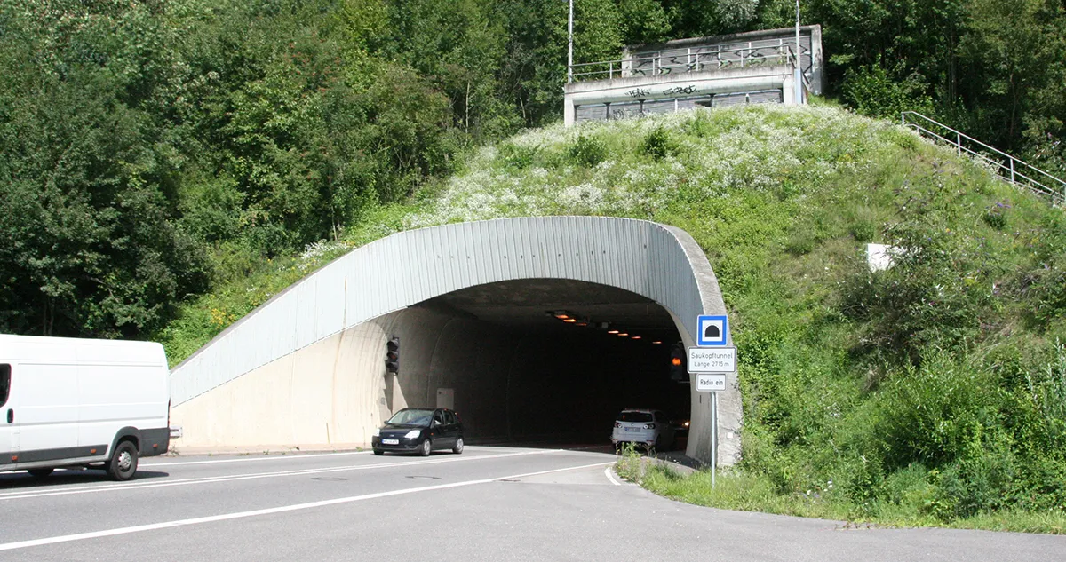 Der Eingang des Saukopftunnels ist in einen bewaldeten Hügel eingelassen. Ein weißer Lieferwagen steht links auf der Straße, während ein dunkles Auto in den Tunnel einfährt. Ein blaues Schild mit Tunnel-Symbol und der Aufschrift „Saukopftunnel Länge 210 m“ steht rechts am Eingang. (KI-Alternativtext)
