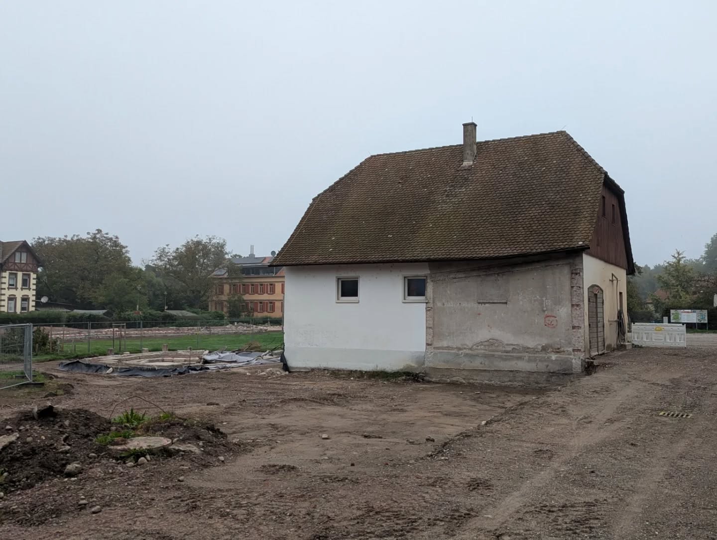 Dieser Alternativtext wurde KI-generiert: Ein Gebäude mit einem schrägen Dach und einer hellen Fassade steht auf einem Grundstück im Umbruch. Im Hintergrund sind weitere Häuser und eine Baustelle zu sehen.