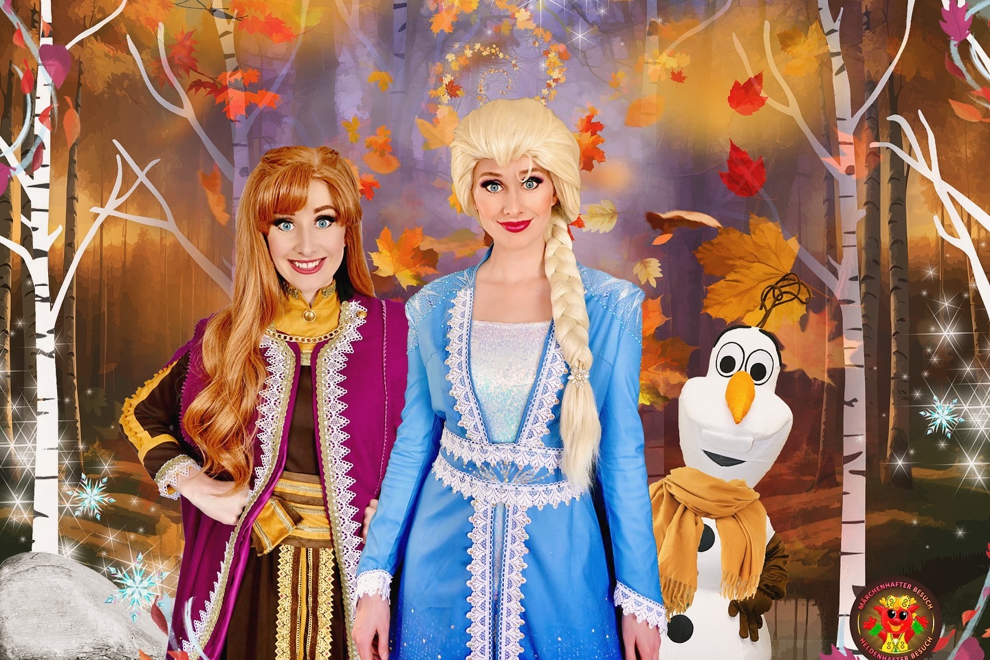 Zwei Personen im Kostüm stehen vor einem herbstlichen Hintergrund mit einem Schneemann. Das Bild ist bunt und fröhlich.